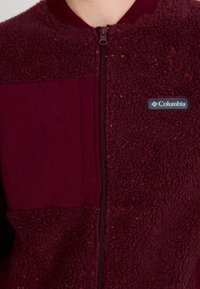 Burgundia fliisjack, millel on tekstureeritud pind, vasakul sujuv paneel ja lukuga sulgemine. Columbia logo on selgelt nähtav.