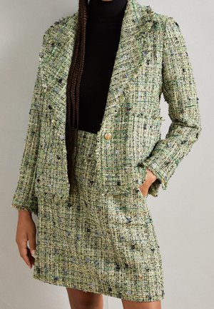 Grüner Tweedblazer und passender Rock kombiniert mit schwarzem Rollkragenpullover, Model mit langen Zöpfen, Hand in der Rocktasche.