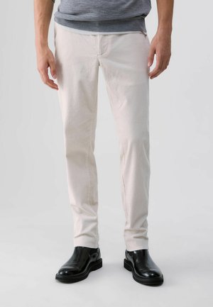 Homme portant un pantalon en velours côtelé blanc et des bottines Chelsea en cuir noir, debout devant un fond uni.