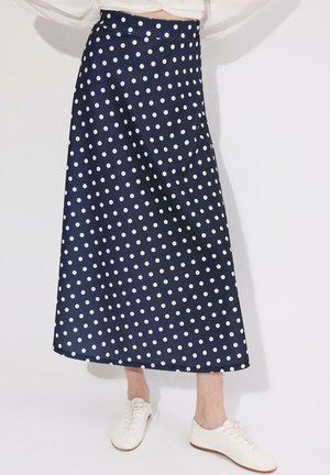 Jupe midi bleu marine à pois blancs portée avec des baskets blanches et un top blanc court, debout devant un fond uni.