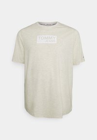 Světle béžové tričko vyrobené z měkké tkaniny s krátkým rukávem. Na přední straně je bílé obdélníkové logo "TOMMY JEANS".