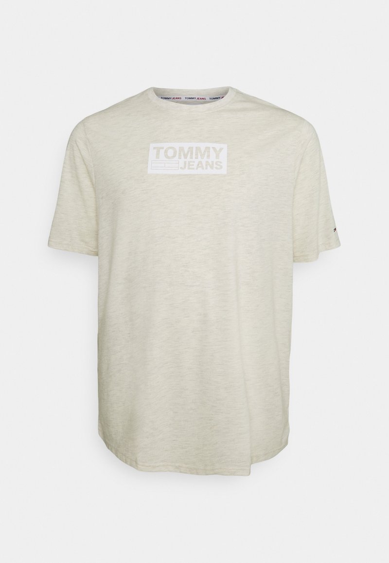 Světle béžové tričko vyrobené z měkké tkaniny s krátkým rukávem. Na přední straně je bílé obdélníkové logo "TOMMY JEANS".
