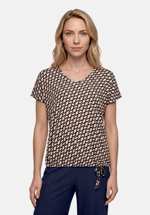 Femme blonde portant un haut à manches courtes à motifs avec un pantalon bleu marine, debout face à un fond clair uni.