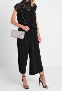 Musta haalaripuku, jossa on pitsinen kaula-aukko ja lyhyet hihat, yhdistettynä hopeatekstuuriseen clutch-laukkuun ja mustiin korollisiin sandaaleihin, joissa on kiiltävää koristetta.