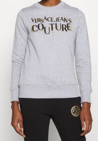 Person wearing a grey sweatshirt with "Versace Jeans Couture" text and black pants with a gold logo patch on the thigh.
