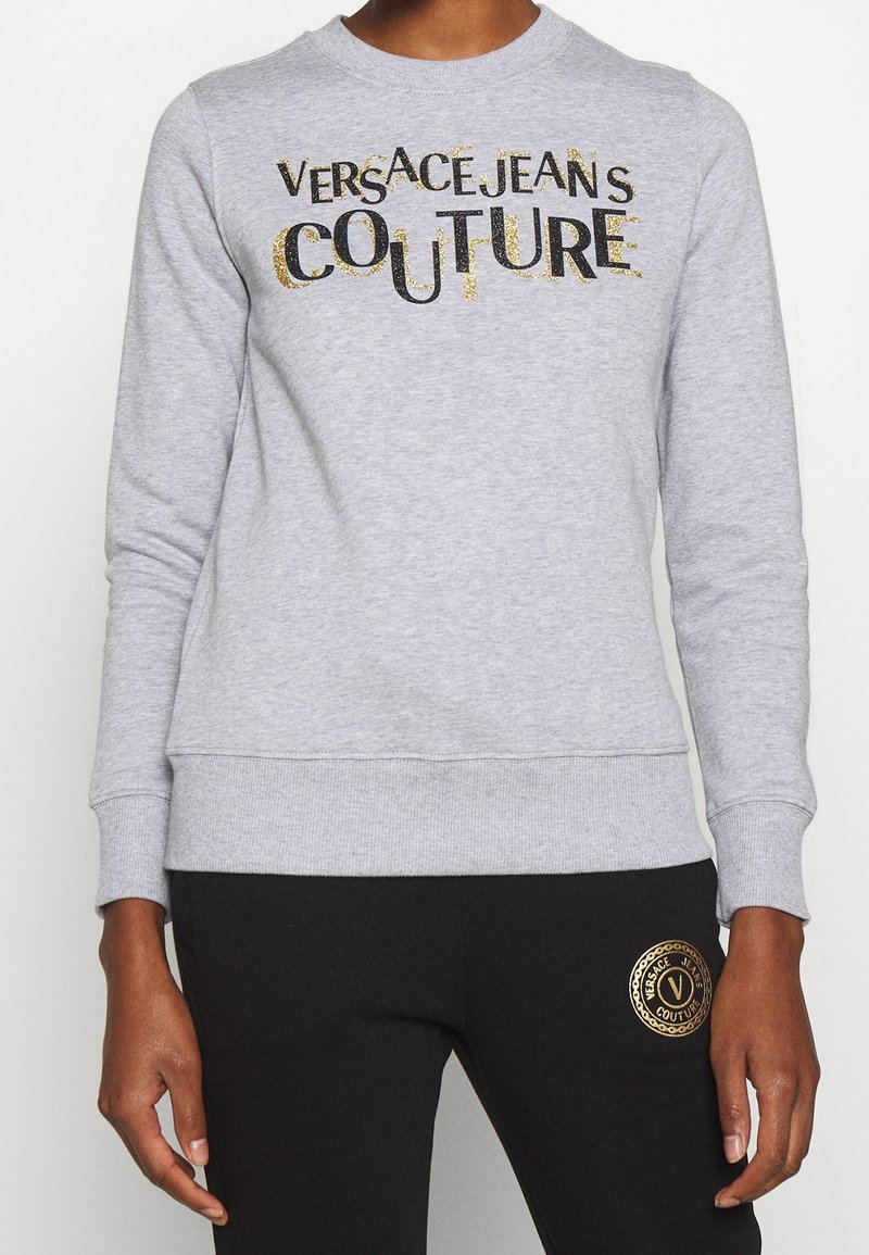 Person wearing a grey sweatshirt with "Versace Jeans Couture" text and black pants with a gold logo patch on the thigh.