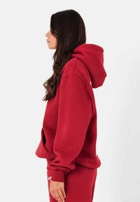 Rode hoodie met een ontspannen pasvorm, grote capuchon en een voorkantzak. De stof lijkt zacht met ribgebreide manchetten en een zoom. Opvallend logo detail aan de voorkant.