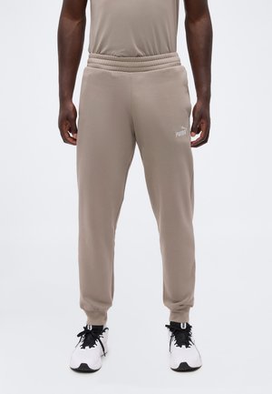 Homme portant un jogger beige Puma et des baskets blanches debout devant un fond uni clair.