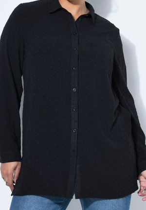 Personne portant une chemise noire texturée à manches longues boutonnée sur un jean bleu, debout contre un mur blanc.