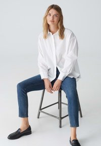 Chemise blanche à boutons, coupe décontractée, manches longues. Portée avec un jean bleu foncé et des mocassins noirs. Tabouret en métal comme siège. Fond simple.