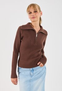 Smeđa rebrasta veste s visokim ovratnikom na patentni zatvarač, koja ima teksturirani uzorak i prilagođen kroj. Uparena s svijetloplavom traper suknjom.