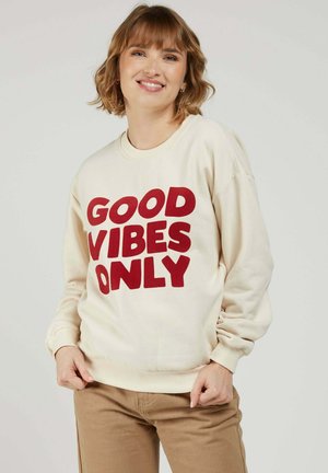 Femme souriante aux cheveux courts bruns portant un sweat-shirt crème avec l'inscription "GOOD VIBES ONLY" en grandes lettres rouges et un pantalon beige.