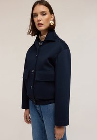 Giacca blu navy con colletto alto, bottoni frontali e due tasche a toppa. Realizzata in tessuto liscio con una vestibilità strutturata e maniche con risvolto.