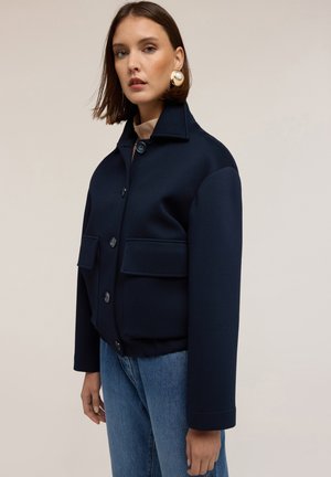 Giacca blu navy con colletto alto, bottoni frontali e due tasche a toppa. Realizzata in tessuto liscio con una vestibilità strutturata e maniche con risvolto.