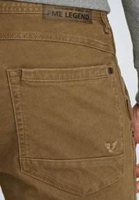 Pantalones cargo marrones con tejido texturizado, que cuentan con un bolsillo trasero y un pequeño logotipo bordado. Detalles de costura y un acento de botón visibles.