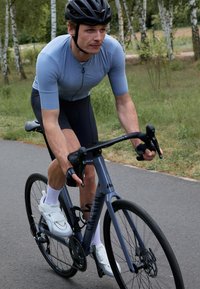 Un cycliste sur un vélo gris porte un maillot bleu clair à manches courtes et un short noir. Le vélo a un cadre aérodynamique et des roues noires.