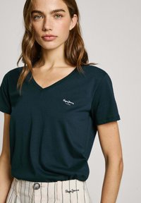 Mörkgrön V-ringad t-shirt i mjukt tyg med en liten vit logotyp på bröstet. Kombinerad med randiga, ljusa byxor.