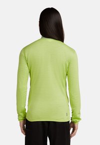 Persoon met lang donker haar, gekleed in een limoen groene lange mouwen shirt en zwarte broek, met de rug naar de camera gericht tegen een effen witte achtergrond.