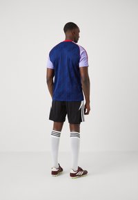 Maglia da calcio di colore blu navy con un motivo geometrico, maniche viola e dettagli rossi; abbinata a pantaloni corti neri e calzettoni a righe bianche.