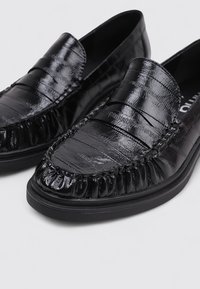 Mokasines negros con un upper de cuero brillante y texturizado, que presentan acentos de diseño plisado y una suela de goma acolchada. Forma de punta redonda.