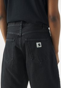 Svarte denimbukser med høy midje, med to baklommer og en firkantet etikett med logo. Diskré tekstur og klassisk snitt.