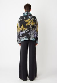 Svetr s multikolorovým květinovým vzorem na černém pozadí, vysokým límcem a oversized střihem, spárovaný s černými širokými kalhotami.