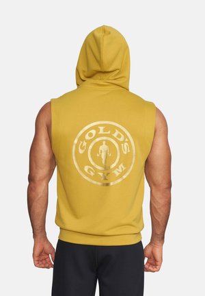 Muskelbepackte Person trägt gelbe ärmellose Kapuzenjacke mit Gold's Gym-Logo auf dem Rücken, steht vor einem schlichten hellen Hintergrund.