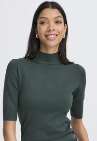Tamnozeleni kratki rukav turtleneck pulover izrađen od glatkog pletenog materijala. Ima prilagođen oblik s rebrastim ovratnikom i manžetama.