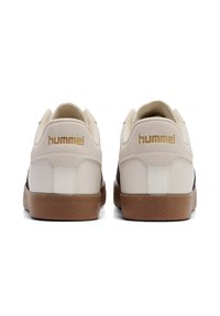 Adidași albi și negri cu o suprafață texturată, talpă din cauciuc bej și logo-ul "hummel" aurit pe călcâi. Design rotund cu accente cusute.