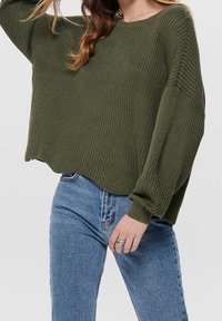 Pull vert olive en tricot lâche avec un large col et des épaules tombantes, associé à un jean en denim bleu délavé. Détail du tissu texturé visible.