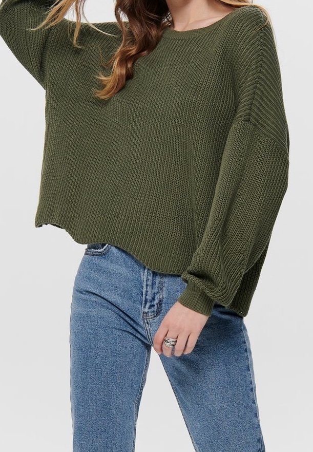 Pull vert olive en tricot lâche avec un large col et des épaules tombantes, associé à un jean en denim bleu délavé. Détail du tissu texturé visible.