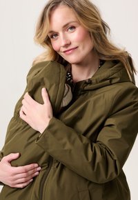 Vrouw in een olijfgroene jas die een baby in de jas houdt, met een beige gebreide muts, terwijl ze vriendelijk naar de camera glimlacht.