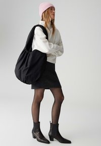 Borsa nera oversize in tessuto morbido, indossata a tracolla, abbinata a una felpa bianca, gonna grigia, calze trasparenti e stivaletti neri.