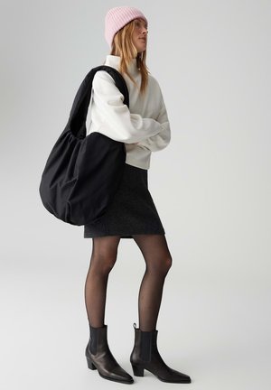 Borsa nera oversize in tessuto morbido, indossata a tracolla, abbinata a una felpa bianca, gonna grigia, calze trasparenti e stivaletti neri.