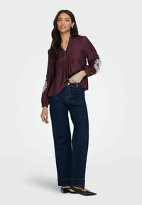 Vinsfarvet bluse med plisseringer og broderede detaljer på ærmerne, parret med mørkeblå brede jeans og sorte flade sko. Glat tekstur, afslappet pasform.