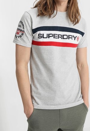 Osoba nosząca szarą koszulkę Superdry z granatowymi i czerwonymi paskami oraz logo na klatce piersiowej i napisem na lewym rękawie.