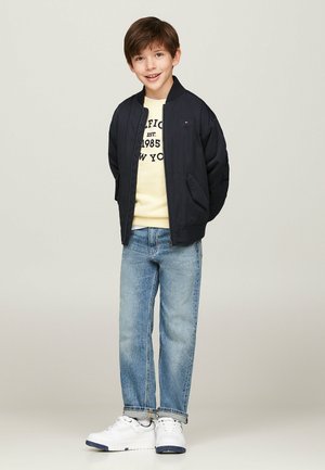 Giacca bomber navy, felpa gialla con grafica, jeans azzurri chiari e sneakers bianche. La giacca presenta una chiusura con zip e tasche laterali.
