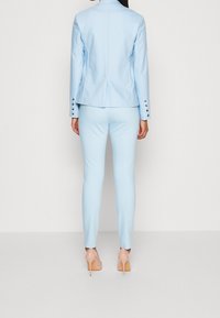 Costume bleu clair sur mesure avec une veste cintrée et un pantalon slim. Tissu lisse, fermeture à un bouton, et boutons décoratifs sur les manches.