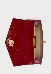 Κόκκινη πατέντα δέρμα τσάντα χειρός με χρυσό λογότυπο, εξαγωνικό σχήμα και μπεζ εσωτερικό. Διαθέτει κλείσιμο με κουμπί και ανάγλυφη υφή.