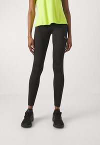Leggings negros de tela elástica con una textura suave, combinados con una camiseta sin mangas de color verde brillante y zapatillas deportivas negras.