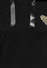 Mustast kangast koos kahe läikiva musta satiinpaela rihmaga ja paremal küljel väikese leopardimustrilise südame detailiga.