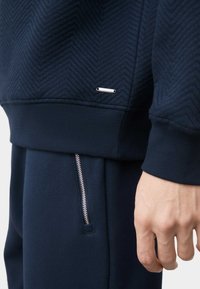 Sudadera azul marino con textura de espiga y puños de canalé; etiqueta de logo metálico en tono plata en el pecho. Pantalones de chándal a juego en azul marino con un bolsillo con cremallera.