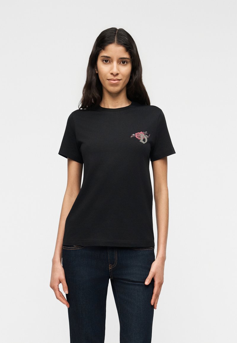 Νεαρή γυναίκα με μακριά σκούρα μαλλιά, ντυμένη με μαύρο μπλουζάκι που έχει ένα μικρό ροζ και γκρι αφηρημένο σχέδιο στο πάνω αριστερό μέρος και σκούρο τζιν.