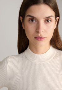 Haut à col roulé en tricot côtelé de couleur blanc cassé, présentant une coupe ajustée et un tissu texturé avec des lignes subtiles verticales.