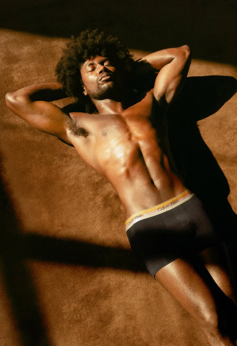 Homme aux cheveux afro allongé torse nu sur une surface marron, les mains derrière la tête, portant des sous-vêtements noirs Calvin Klein, baigné d’une lumière solaire chaude.