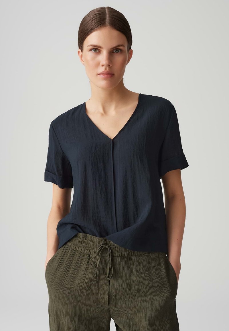 Top blu scuro a maniche corte con scollatura a V, realizzato in tessuto testurizzato. Abbinato a pantaloni plissettati di un verde chiaro con vita elasticizzata.
