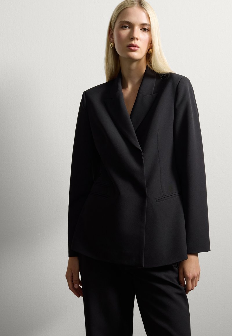 Joven mujer con cabello rubio largo viste una chaqueta negra a medida y pantalones a juego, de pie contra una pared clara y lisa.