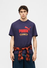 Νέος άντρας που φοράει μπλε μπλουζάκι Puma King και έχει δεμένο κόκκινο-μαύρο καρό πουκάμισο γύρω από τη μέση του, στέκεται μπροστά σε λευκό φόντο.