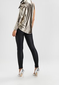 Metallic gold long-sleeve top with shoulder ruffles paired with black skinny pants and silver high-heeled shoes. Smooth texture and fitted design.