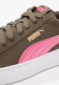 Zamszowy but sportowy w kolorze oliwkowej zieleni z różowymi błyszczącymi akcentami, teksturowanymi sznurowadłami, złotym logo "PUMA" i białą gumową podeszwą.